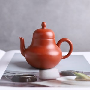 May include: A small, reddish-brown ceramic teapot with a rounded body, curved spout, and a loop handle. The lid has a small knob on top. The teapot is sitting on a white surface.
