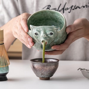 May include: A light green ceramic bowl shaped like a creature with a spout pouring green matcha tea into a dark metallic bowl. Beside it sits a bamboo matcha whisk on a matching green and black stand, and a small metal tea strainer nearby.