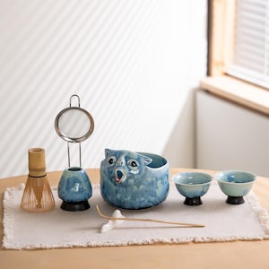 Puede incluir: Un juego de té matcha que incluye un batidor de bambú, un colador de metal, un soporte de cerámica azul para el batidor, un cuenco de cerámica azul con forma de gato con la boca abierta, dos tazas pequeñas de cerámica azul y una cuchara de bambú.