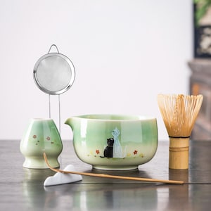 Puede incluir: Un juego de té matcha con un cuenco de cerámica verde claro con una ilustración de gato, un batidor de bambú, una cuchara de té, una taza de té de cerámica, un colador de acero inoxidable y un soporte de cuchara de té de cerámica blanca.