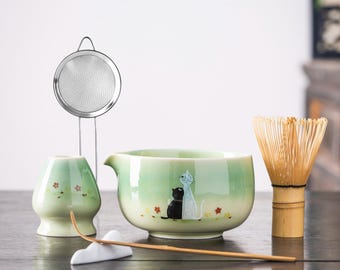 Puede incluir: Un juego de té matcha con un cuenco de cerámica verde claro con una ilustración de gato, un batidor de bambú, una cuchara de té, una taza de té de cerámica, un colador de acero inoxidable y un soporte de cuchara de té de cerámica blanca.