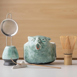 May include: A matcha tea set featuring a light green glazed ceramic bowl shaped like a creature with an open mouth, a matching green ceramic whisk holder with a metal mesh strainer, a bamboo whisk, and a thin bamboo scoop resting on a white stand.