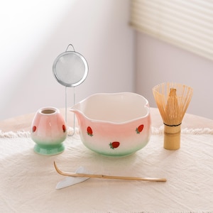 Op de afbeelding: Een matcha theeset met een roze en groene keramische kom en accessoires. De kom is versierd met aardbeien. Inclusief een bamboe klopper, een theelepel, een thee zeef en een keramische klopperhouder.