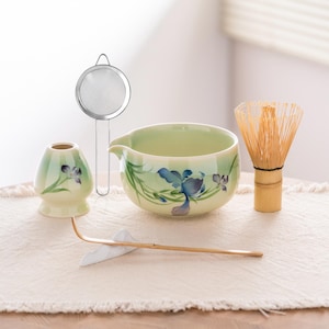 May include: A matcha tea set comprising a light green ceramic bowl and whisk, a small ceramic container, a bamboo scoop, and a stainless steel strainer. The bowl and container are decorated with blue floral designs. The whisk is made of bamboo.
