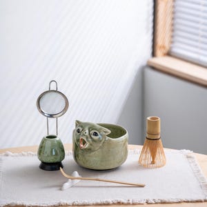 May include: A matcha tea set featuring a light green ceramic bowl shaped like a cat with an open mouth, a matching green whisk holder with a metal mesh sieve, a bamboo whisk, and a long bamboo tea scoop resting on a small white ceramic stand.