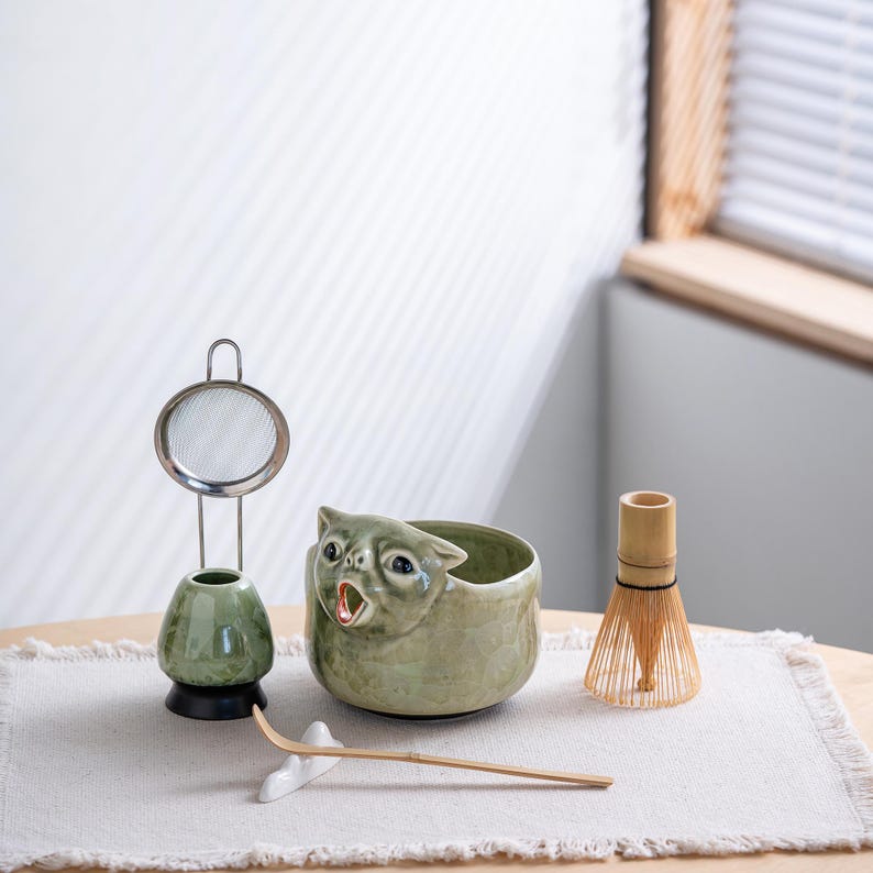 May include: A matcha tea set featuring a light green ceramic bowl shaped like a cat with an open mouth, a matching green whisk holder with a metal mesh sieve, a bamboo whisk, and a long bamboo tea scoop resting on a small white ceramic stand.