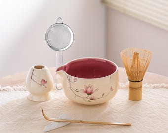 May include: A matcha tea set featuring a cream-colored ceramic bowl with a maroon interior, a bamboo whisk, a small ceramic container, a bamboo scoop, and a stainless steel strainer. The set is decorated with pink floral designs.