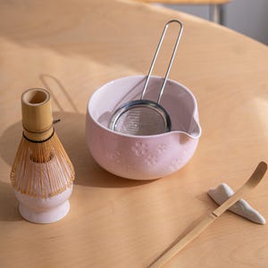 May include: A matcha tea set featuring a light pink ceramic bowl with floral designs, a stainless steel strainer, a bamboo whisk, and a bamboo scoop. The set is arranged on a light wooden surface.