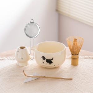 May include: A matcha tea set with a cream-colored bowl featuring a black and white dog illustration. The set includes a bamboo whisk, a ceramic tea scoop rest, a tea caddy, and a stainless steel strainer. The items are arranged on a beige cloth.