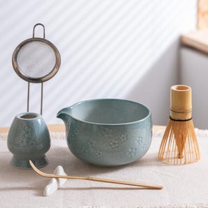 May include: A matcha tea set with a celadon-coloured bowl, whisk, and ceramic accessories. The bowl and accessories are decorated with a floral pattern. A bamboo whisk and a metal tea strainer are also included.