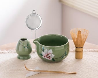 May include: A matcha tea set featuring a green ceramic bowl with a floral design, a bamboo whisk, a small ceramic container, a stainless steel strainer, and a bamboo scoop. The set is arranged on a neutral-colored surface.