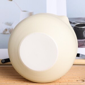 Puede incluir: Un cuenco de cer&aacute;mica de color crema con una base interior blanca. El cuenco tiene forma redondeada con un peque&ntilde;o pico en el lateral. El cuenco est&aacute; sobre una superficie de madera.