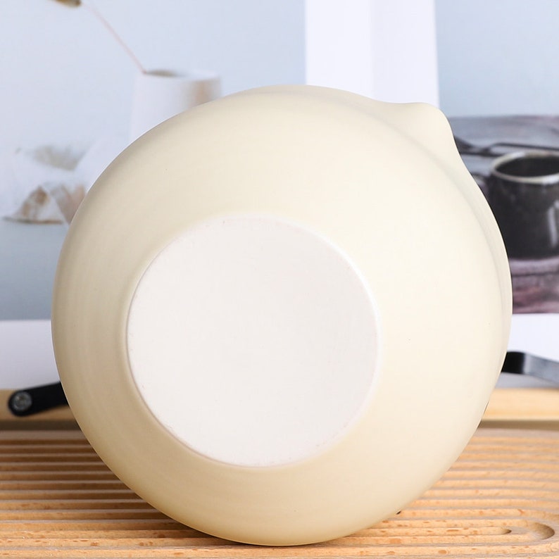 Puede incluir: Un cuenco de cer&aacute;mica de color crema con una base interior blanca. El cuenco tiene forma redondeada con un peque&ntilde;o pico en el lateral. El cuenco est&aacute; sobre una superficie de madera.