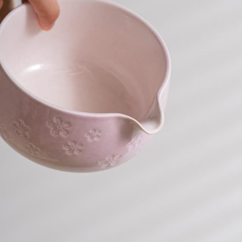 May include: A light pink ceramic mixing bowl featuring a smooth interior and a molded cherry blossom floral pattern on the exterior. The bowl includes a small, integrated pouring spout on the rim for controlled liquid transfer.