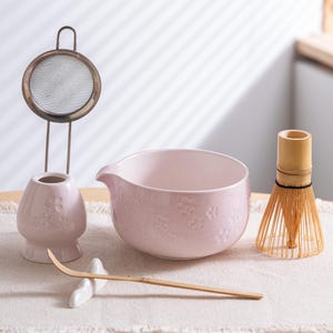 May include: A matcha tea set featuring a light pink ceramic bowl with floral detailing, a bamboo whisk, a whisk holder, a sifter, and a bamboo scoop. The set is arranged on a white surface.
