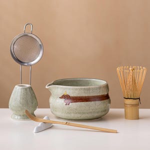 Op de afbeelding: Een matcha-theeset bestaande uit een lichtgroene gespikkelde keramische kom met een bruine teckel-illustratie, een bijpassende gardehouder met metalen zeef, een bamboe garde en een lange bamboe theeschep op een kleine witte standaard.