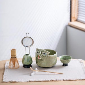 May include: A matcha tea set featuring a light green ceramic bowl shaped like a cat with an open mouth, a bamboo whisk, a whisk holder, a metal mesh strainer, a small green ceramic cup on a black base, and a long bamboo scoop on a white rest.