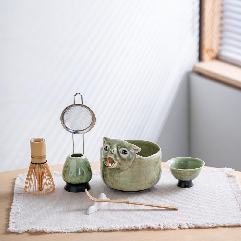 May include: A matcha tea set featuring a light green ceramic bowl shaped like a cat with an open mouth, a bamboo whisk, a whisk holder, a metal mesh strainer, a small green ceramic cup on a black base, and a long bamboo scoop on a white rest.