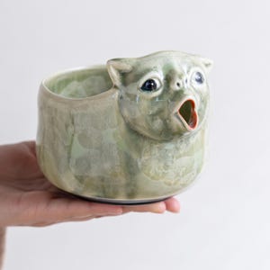 May include: A ceramic bowl featuring a sculpted animal head with a wide, open mouth. The bowl has a glossy, pale green glaze with subtle crystalline patterns throughout the surface. The animal face includes dark, round eyes and pointed ears.
