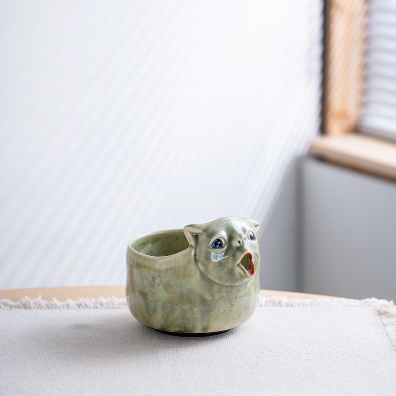 May include: A ceramic creamer shaped like a cat with a light green glazed finish. The cat figure features blue eyes and an open mouth that serves as the pouring spout. The vessel has a rounded body and a smooth, glossy texture throughout the surface.