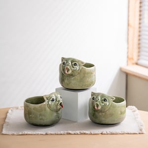 May include: Three sage green ceramic bowls shaped like cat heads with wide-open mouths. Each bowl features a glossy, mottled glaze finish, dark eyes, and pointed ears. One bowl is elevated on a white block, while two sit on the surface below.