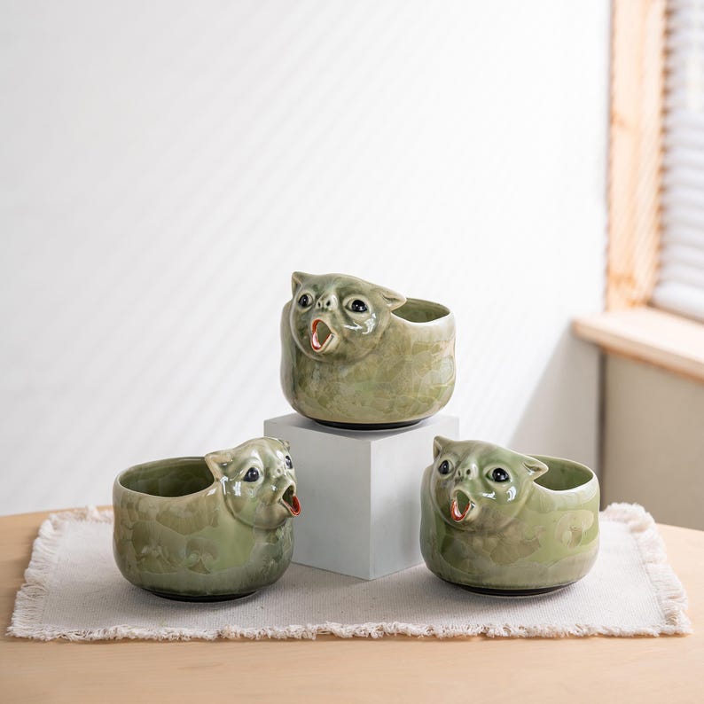 May include: Three sage green ceramic bowls shaped like cat heads with wide-open mouths. Each bowl features a glossy, mottled glaze finish, dark eyes, and pointed ears. One bowl is elevated on a white block, while two sit on the surface below.
