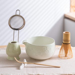 Puede incluir: Un juego de té matcha con un cuenco de cerámica verde claro con diseños florales, un batidor de bambú, una cuchara de bambú, un soporte para batidor de cerámica y un colador de té de acero inoxidable. El juego está dispuesto sobre un paño de color neutro.
