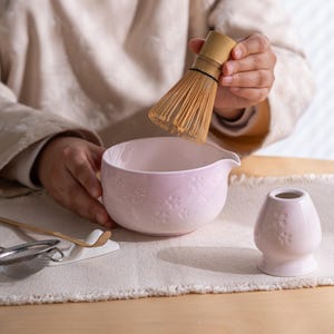 May include: A light pink ceramic matcha bowl with embossed floral designs, a bamboo whisk, and a small matching ceramic stand. The bowl has a pouring spout. The whisk is held over the bowl, suggesting the preparation of matcha tea.