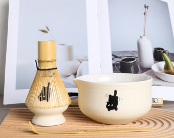 May include: A matcha tea set featuring a bamboo whisk, a cream-colored bowl with a black graphic, and a bamboo scoop. The whisk is light brown with a white base. The bowl has a curved spout. The set is on a wooden tray.