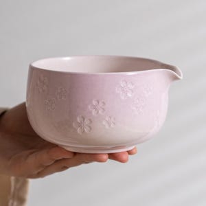 May include: A light pink ceramic bowl featuring a subtle embossed cherry blossom pattern across the exterior surface. The bowl has a smooth, rounded shape with a small, integrated pouring spout on the right side of the rim.