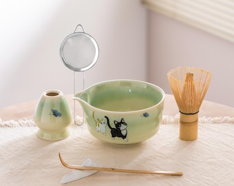 May include: A matcha tea set featuring a light green ceramic bowl with cat illustrations, a bamboo whisk, a tea scoop, a tea caddy, and a stainless steel strainer. The set is arranged on a beige surface.