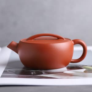 May include: A small, round, reddish-brown teapot with a curved handle and a lid with a loop. The teapot has a spout and sits on a white surface. The teapot is likely made of ceramic.