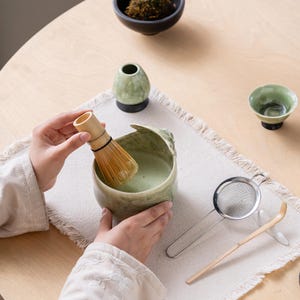 May include: A light green ceramic matcha bowl containing green tea, a bamboo whisk, a stainless steel mesh strainer, a bamboo tea scoop, a small green ceramic tea cup, and a green whisk holder. The items are arranged for preparing matcha tea.