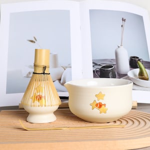 Puede incluir: Un juego de t&eacute; matcha con un batidor de bamb&uacute;, un cuenco color crema con ilustraciones de peces y estrellas y una cuchara de bamb&uacute;. El batidor y el cuenco est&aacute;n sobre una bandeja de madera, con un libro al fondo.