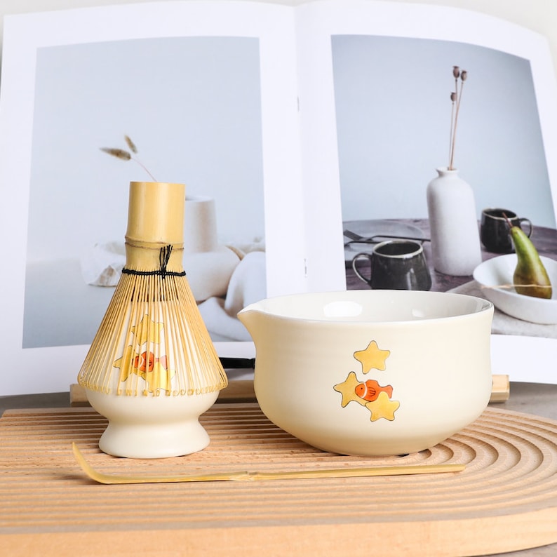 Puede incluir: Un juego de t&eacute; matcha con un batidor de bamb&uacute;, un cuenco color crema con ilustraciones de peces y estrellas y una cuchara de bamb&uacute;. El batidor y el cuenco est&aacute;n sobre una bandeja de madera, con un libro al fondo.