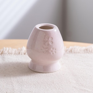 May include: A small, light pink ceramic vase with a rounded body and a flared base. The vase features three embossed flower designs on the front. The opening is circular. The vase is sitting on a cream-colored surface.
