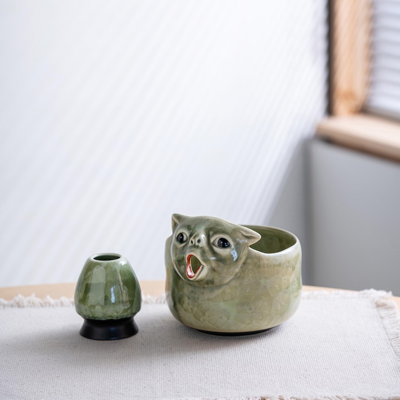 May include: A sage green ceramic matcha bowl featuring a sculpted cat face with an open mouth, paired with a matching sage green ceramic whisk holder that has a solid black base. Both items have a glossy, mottled glaze finish.
