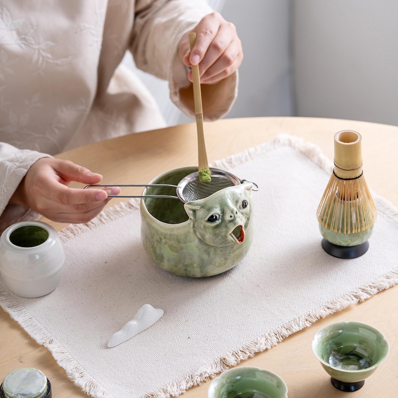 May include: A light green ceramic bowl shaped like a cat head with an open mouth, a metal mesh strainer, a bamboo scoop with green powder, a bamboo whisk on a stand, and small matching ceramic cups for preparing and serving matcha tea.