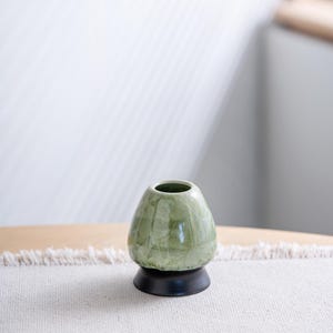 May include: A ceramic matcha whisk holder featuring a glossy, light sage green glaze with a crystalline pattern. The holder has a rounded, tapered body shape and sits on a solid, dark brown circular base, providing a stable foundation for the tool.