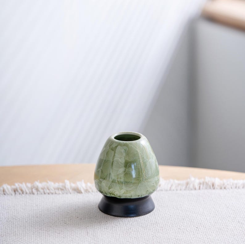 May include: A ceramic matcha whisk holder featuring a glossy, light sage green glaze with a crystalline pattern. The holder has a rounded, tapered body shape and sits on a solid, dark brown circular base, providing a stable foundation for the tool.