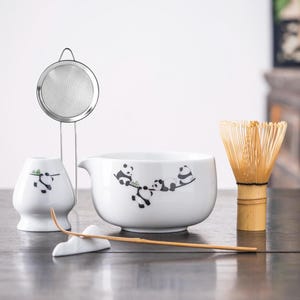 May include: A white ceramic tea set with a panda bear design. The set includes a bowl, whisk, strainer, and a small holder. The bamboo whisk is light brown. The panda design is black and white.