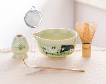 May include: A matcha tea set featuring a light green ceramic bowl with cat illustrations, a bamboo whisk, a tea scoop, a tea strainer, and a ceramic tea container. The set is arranged on a cream-colored cloth.