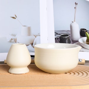 Puede incluir: Un juego de cuenco y batidor de matcha de color crema. El cuenco tiene forma redondeada con un pico, y el batidor tiene una cabeza bulbosa. El juego se exhibe sobre una superficie de madera, con un fondo neutro.