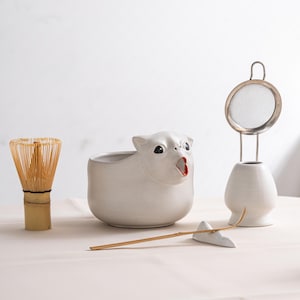 Op de afbeelding: Een verzameling witte keramische theeceremonie-artikelen. Inclusief een klopper, een zeef, een kleine beer-vormige kom met een rode binnenkant, een bamboe lepel en een keramische lepelsteun. De items zijn gerangschikt op een lichtgekleurd oppervlak.