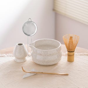May include: A matcha tea set comprising a ceramic bowl, whisk, whisk holder, scoop, and a stainless steel strainer. The ceramic pieces have a speckled finish in neutral tones. The bamboo whisk is light brown.