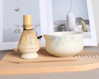 May include: A matcha tea set featuring a bamboo whisk, a ceramic bowl with blue floral designs, and a bamboo scoop. The bowl is cream-colored and has a small spout. The whisk is light brown and has a black string around the handle.