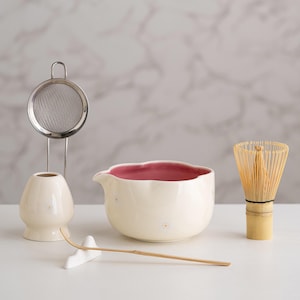 May include: A matcha tea set featuring a cream-colored ceramic bowl with a pink interior, a bamboo whisk, a cream-colored ceramic tea bowl holder, a stainless steel strainer, and a bamboo scoop. The set is designed for preparing matcha tea.