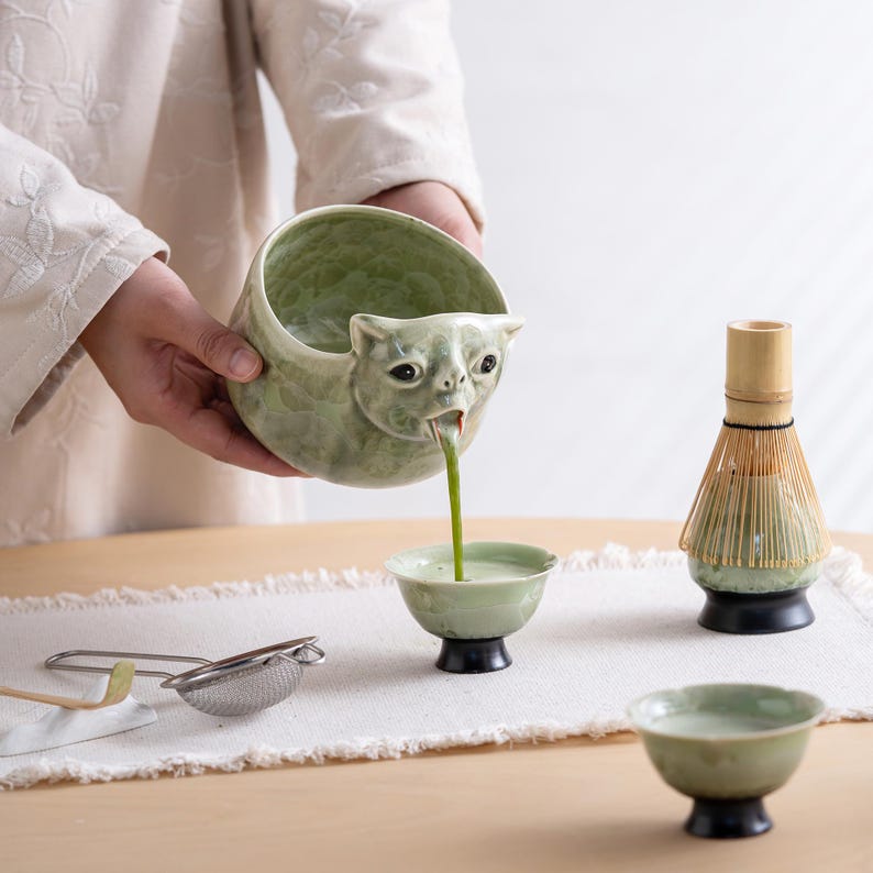May include: A light green ceramic bowl shaped like a cat head pouring matcha into a small matching cup. A bamboo whisk on a black stand, a metal mesh strainer, and an additional small green ceramic cup are arranged on a white fabric runner.