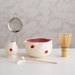 Könnte beinhalten: Ein Matcha-Teeset mit einer cremefarbenen Keramikschale mit rosa Innenraum und Erdbeermotiven, einem Bambusbesen, einem Besenhalter, einem Edelstahl-Sieb und einem Bambuslöffel. Das Set ist für die Zubereitung von Matcha-Tee konzipiert.