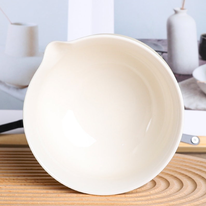 Puede incluir: Un cuenco de cer&aacute;mica color crema con un pico vertedor. El cuenco tiene un acabado liso y brillante y una forma redondeada. El cuenco est&aacute; sobre una superficie de madera.
